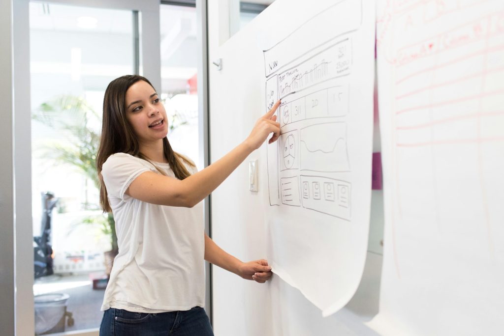 female in front of meeting at whiteboard, pointing to diagram